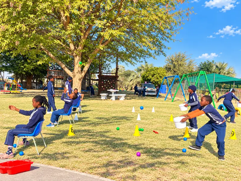 Children playing a catching game with buckets, cones and coloured balls under a big tree.