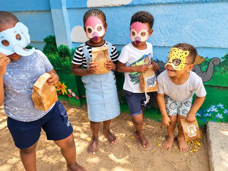 Four children in paper animal masks holding party bags beside a painted wall.